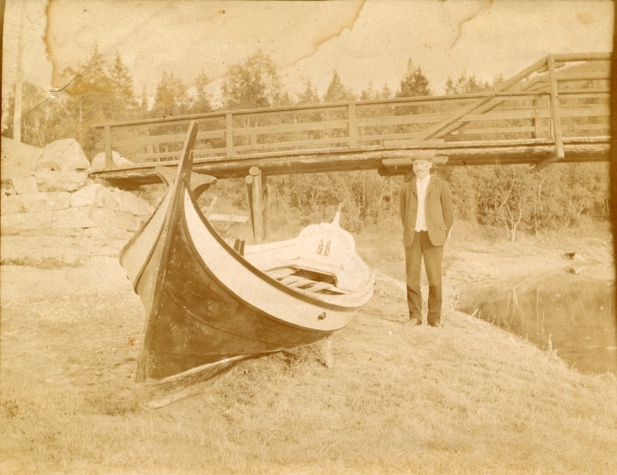 Portrett. Mann står foran bru ved siden av en nordlandsbåt. Karsten Venes med færing. Mobekken og Mobekkbrua.
