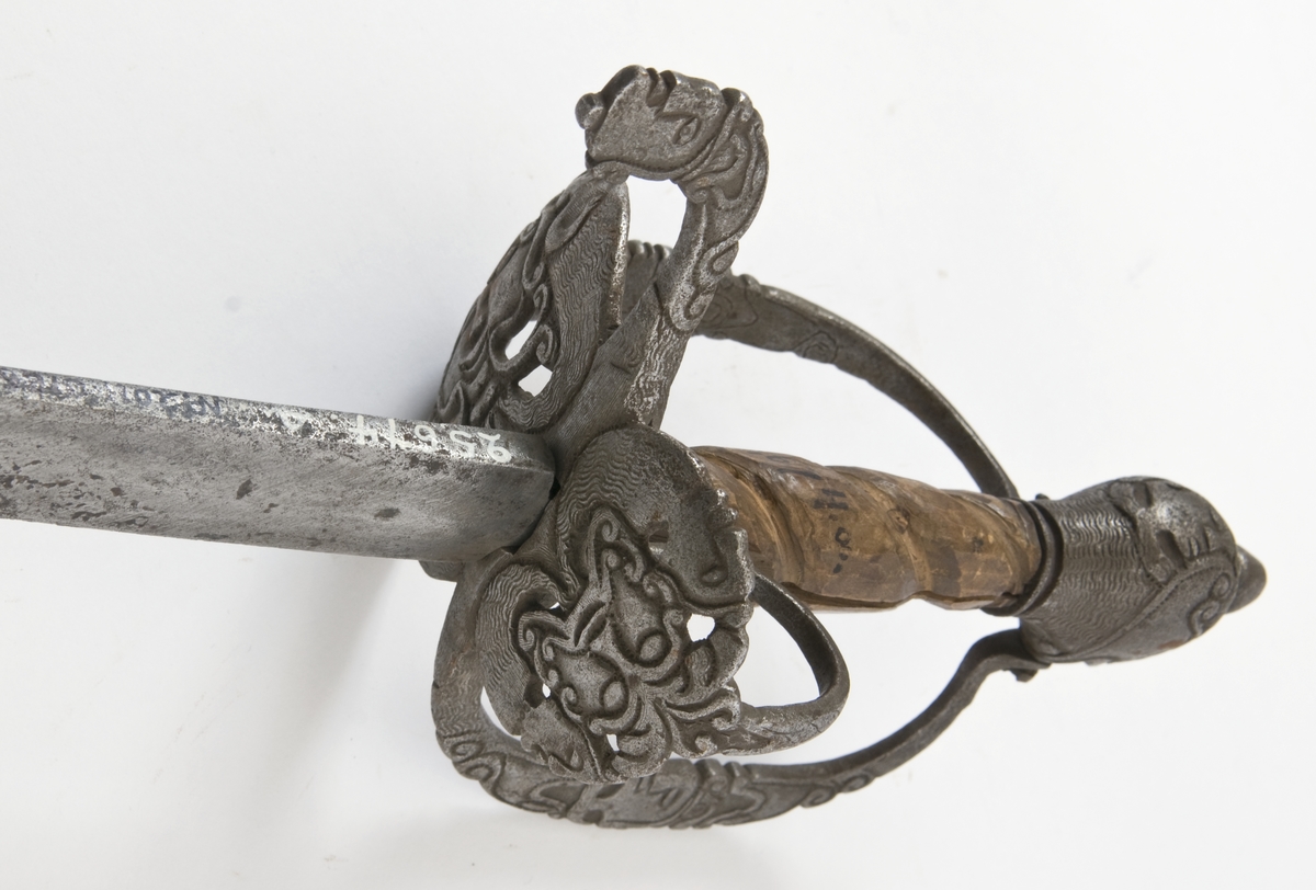 Hundehodeformet knapp med nitteknapp og skåret dekor.Tregrep  S formede parerstenger med fortykninger på endene.Fremre parerstang går over i håndbøylen, som er skrudd til knappen.Rund perforet stikkplate på utsiden med sidebøyle som er skrudd til knappen.Midere perforert stikkplate på innsiden med tombøyle. Hele festet har rikt skåret dekor bl a med masker.
Eneegget, krum klinge med kileformet tverrsnitt og ryggslipning mot spissen..