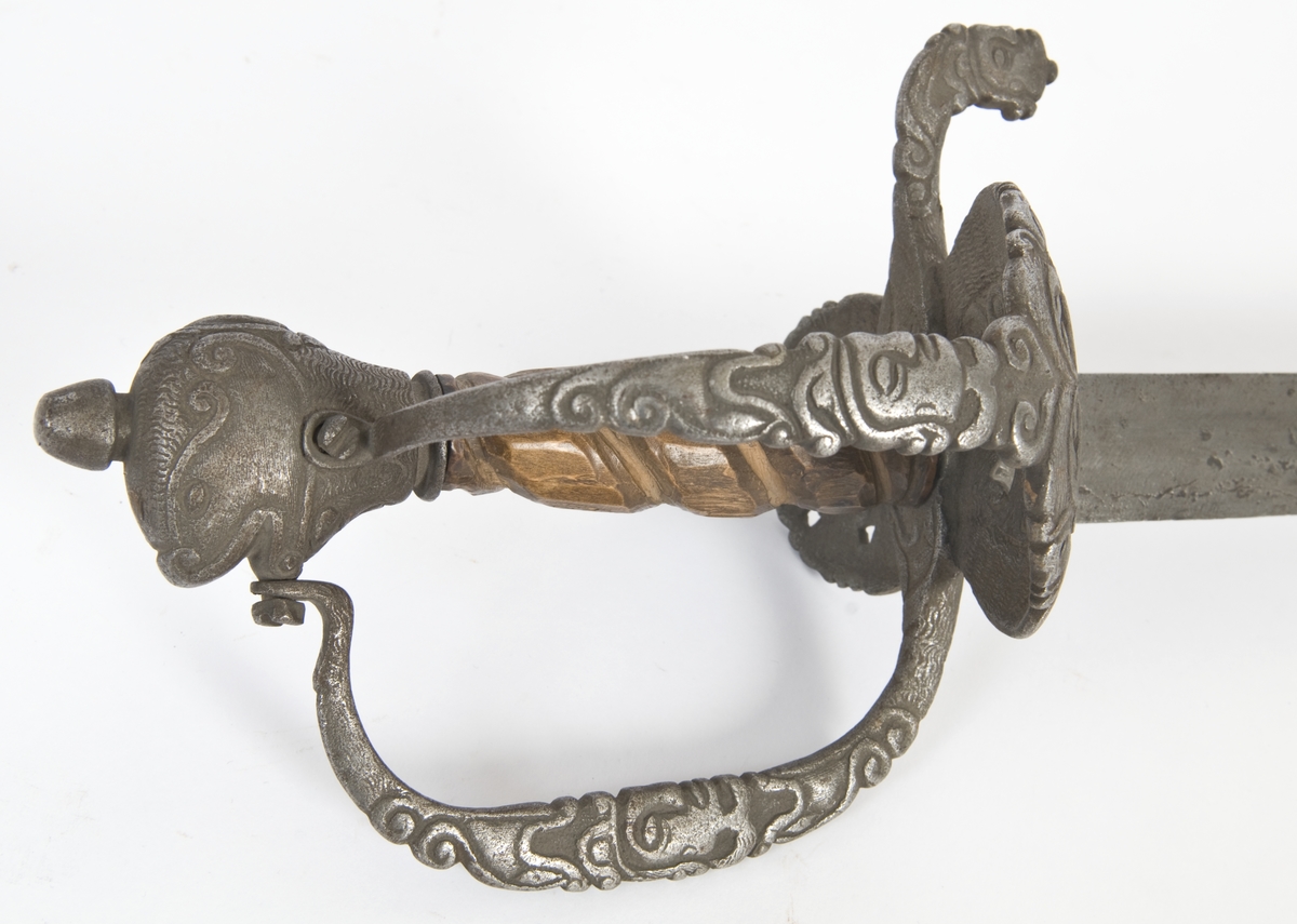 Hundehodeformet knapp med nitteknapp og skåret dekor.Tregrep  S formede parerstenger med fortykninger på endene.Fremre parerstang går over i håndbøylen, som er skrudd til knappen.Rund perforet stikkplate på utsiden med sidebøyle som er skrudd til knappen.Midere perforert stikkplate på innsiden med tombøyle. Hele festet har rikt skåret dekor bl a med masker.
Eneegget, krum klinge med kileformet tverrsnitt og ryggslipning mot spissen..