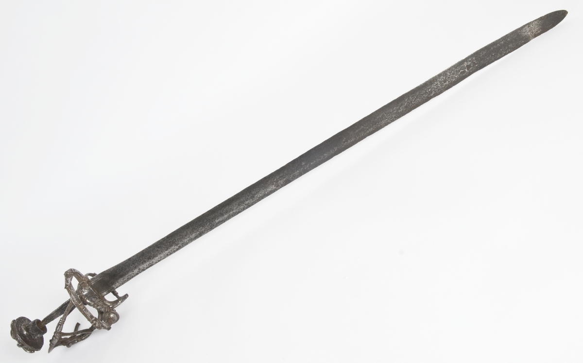 Soppformet knapp med nitteknapp Oversiden av knappen har skåret og meislet dekor. Grep mangler .Asymmetrisk  feste av runde bøyler Sidering på utsiden som ender i en knapp. Sideringen på innsiden er borte. Klingebrekker på innsiden. Håndbøylen er brukket rett under knappen Sidebøyle og tersbøyle. Bøylene har smidd og meislet dekor. Flere bøyler på innsiden er brukket.
Rett tveegget klinge med ovalt tverrsnitt  Klingen merket mes en snal bølgelinje innlagt i kobber.