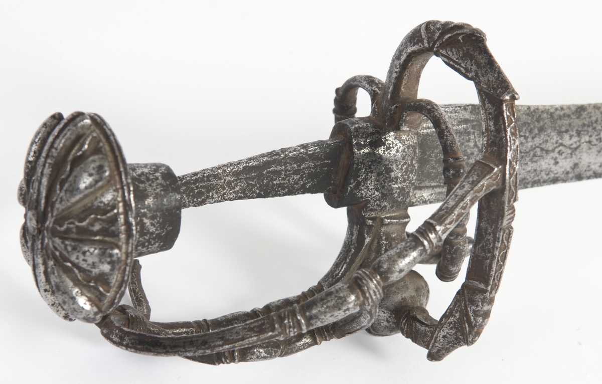 Soppformet knapp med nitteknapp Oversiden av knappen har skåret og meislet dekor. Grep mangler .Asymmetrisk  feste av runde bøyler Sidering på utsiden som ender i en knapp. Sideringen på innsiden er borte. Klingebrekker på innsiden. Håndbøylen er brukket rett under knappen Sidebøyle og tersbøyle. Bøylene har smidd og meislet dekor. Flere bøyler på innsiden er brukket.
Rett tveegget klinge med ovalt tverrsnitt  Klingen merket mes en snal bølgelinje innlagt i kobber.