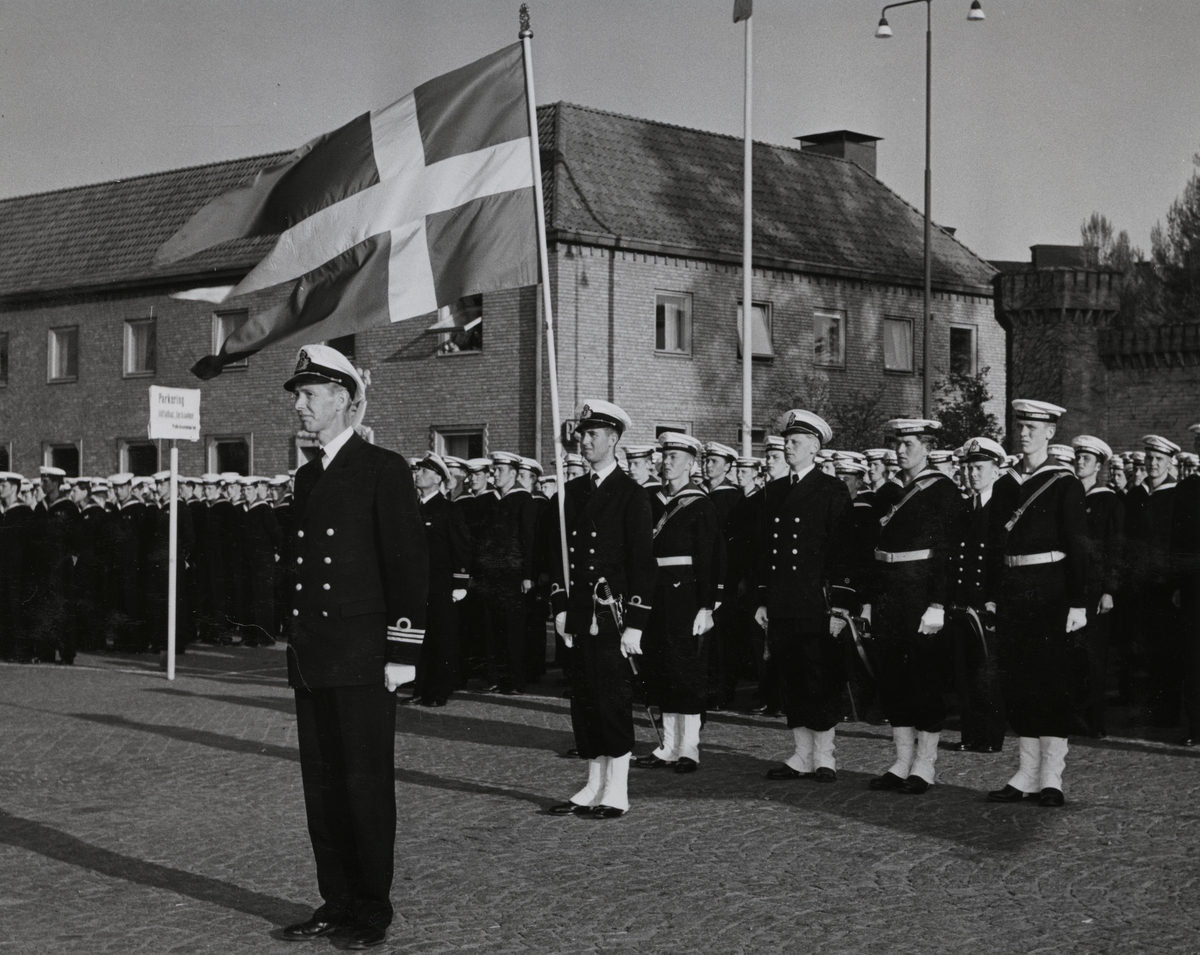 En grupp sjömän står givakt på stortorget. En fänrik i framledet håller i en stor, svensk tretungad flagga. Framför honom står en kapten. I bakgrunden syns det gamla posthuset på adressen Stortorget 3.