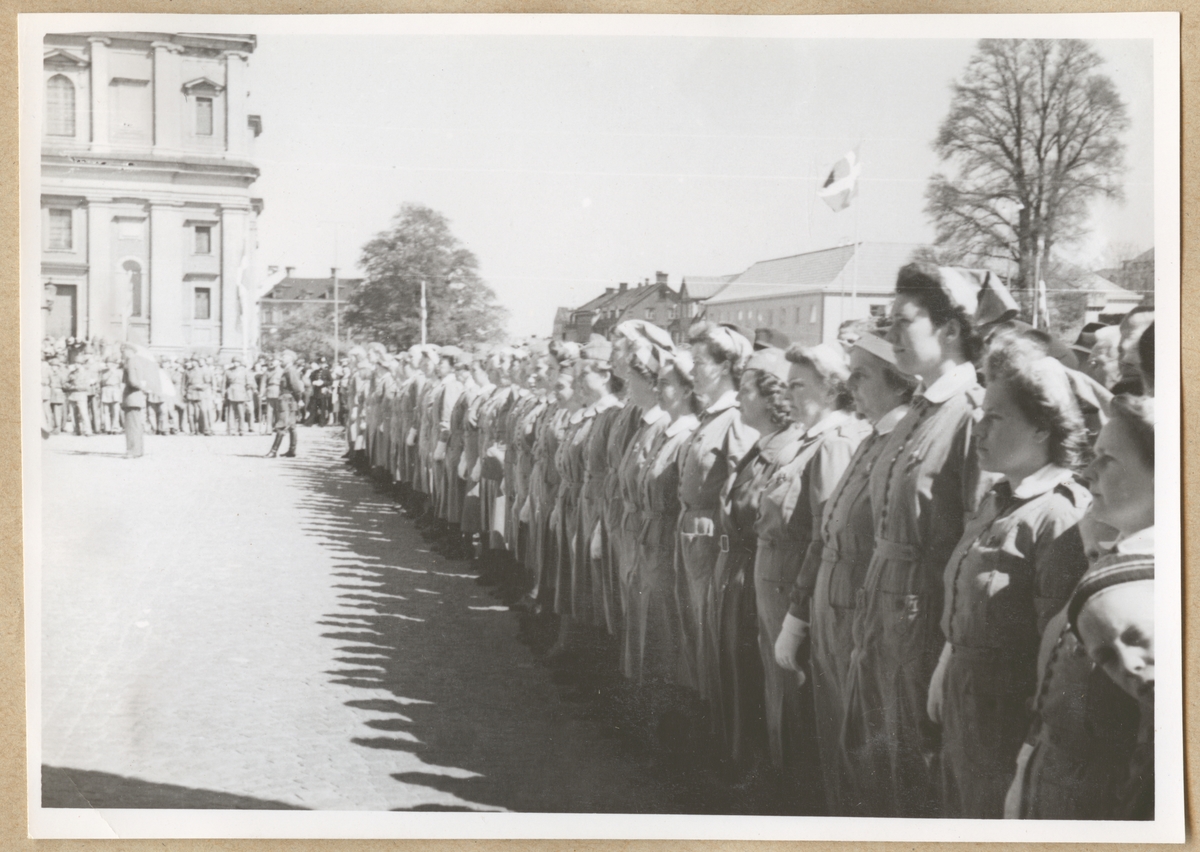 Uniformsklädda lottor uppställda på rad på stortorget i Karlskrona. I bakgrunden skymtar gamla posthuset och Fredrikskyrkan. Framför kyrkan står en grupp soldater.