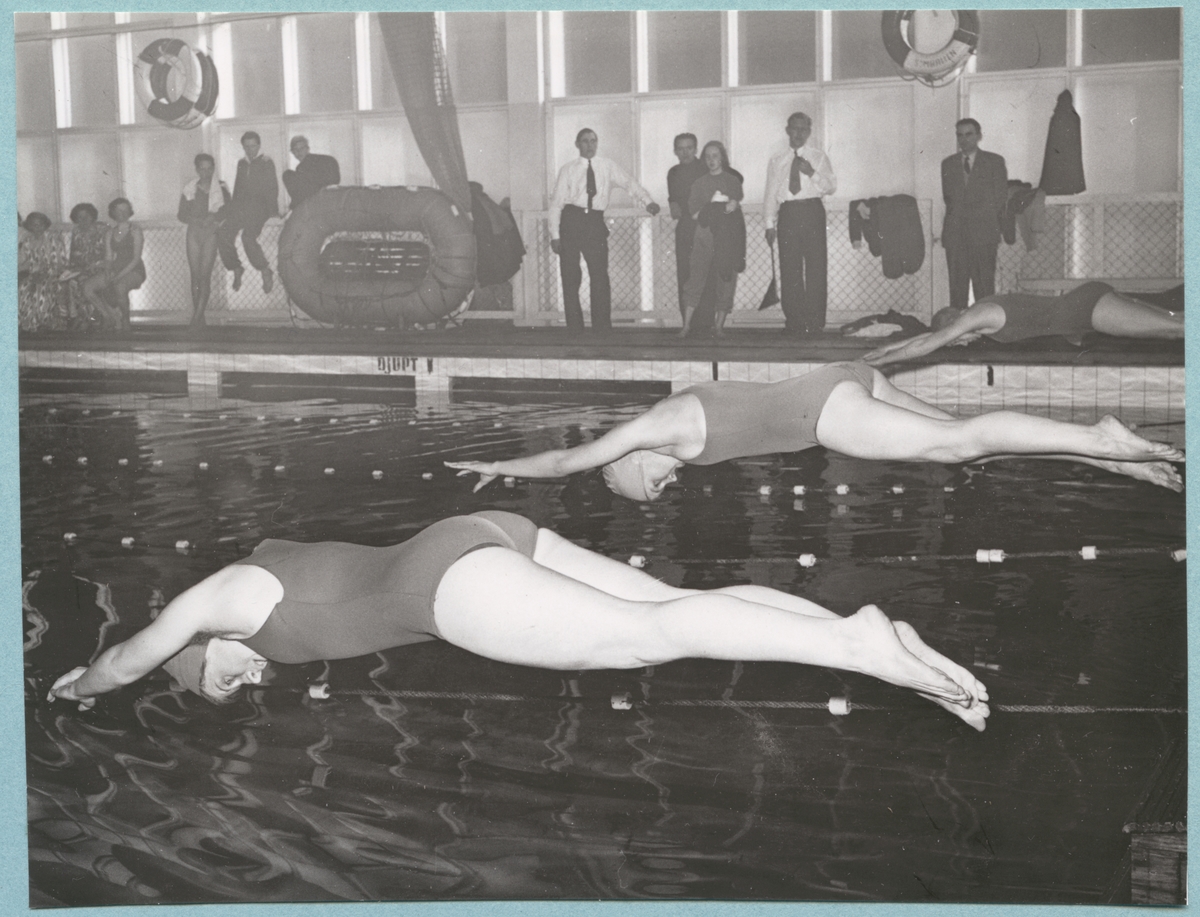 Tre kvinnliga simmare dyker ner i simbassängen tillhörande Sparres simhall. Alla tre är klädda i baddräkter och badmössor. Längs med simbassängens kant står och sitter åskådare. På bassängkanten står även en liten livbåt och på väggen hänger livbojar. Fotot är daterat till 15-3-1952.