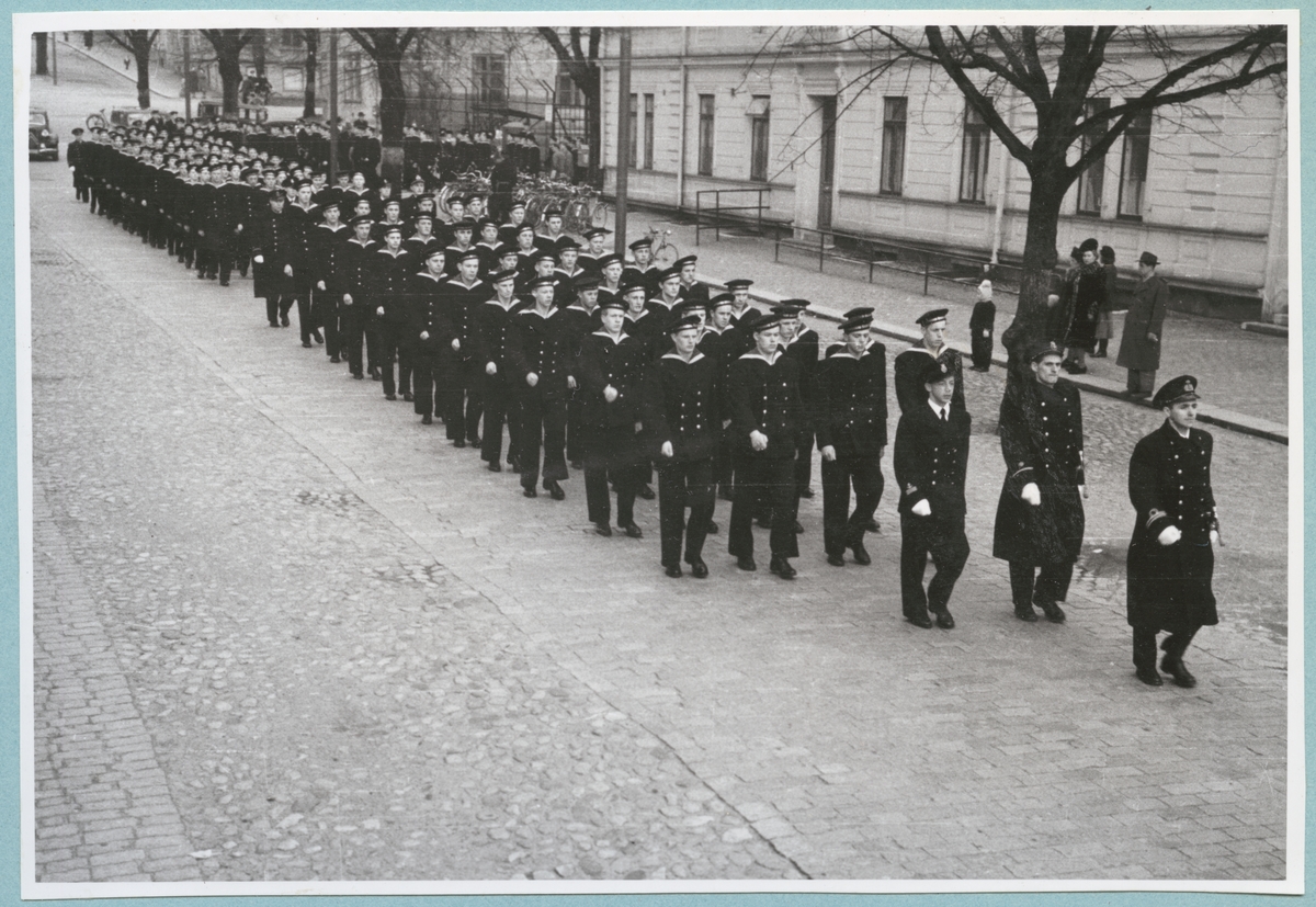 Uniformsklädda flottister marscherar längs med Drottninggatan under ledning av en löjtnant. Den stenlagda gatan kantas av bataljon Sparres kanslibyggnad. På gatan står även ett par civila åskådare.
Kyrkparad för nyinryckta värnpliktiga.