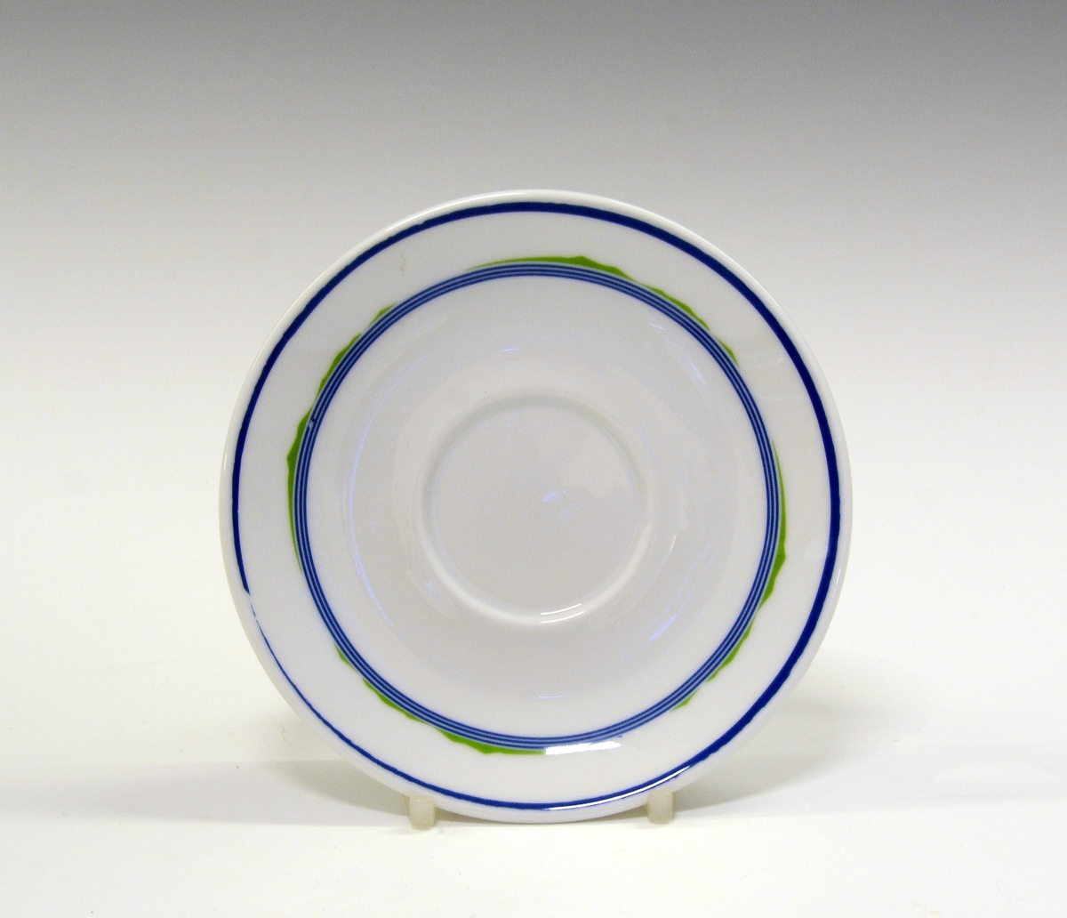 Kaffeskål av porselen. Hvit glasur. Dekorert med blå stripe ytterst på fanen og tre smale blå striper med noe grønt på innerst på fanen.
Modell: Roma