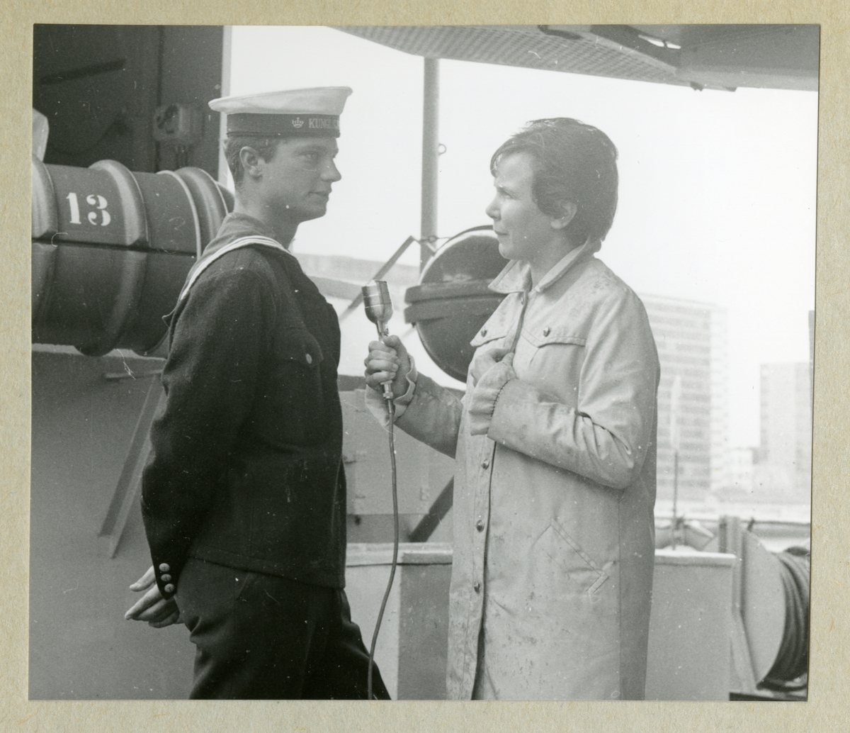 Bilden föreställer en kvinna med en mikrofon som intervjuar kronprinsen Carl Gustaf, som står klädd i uniform. Bilden är tagen under minfartyget Älvsnabbens långresa 1966-1967.
