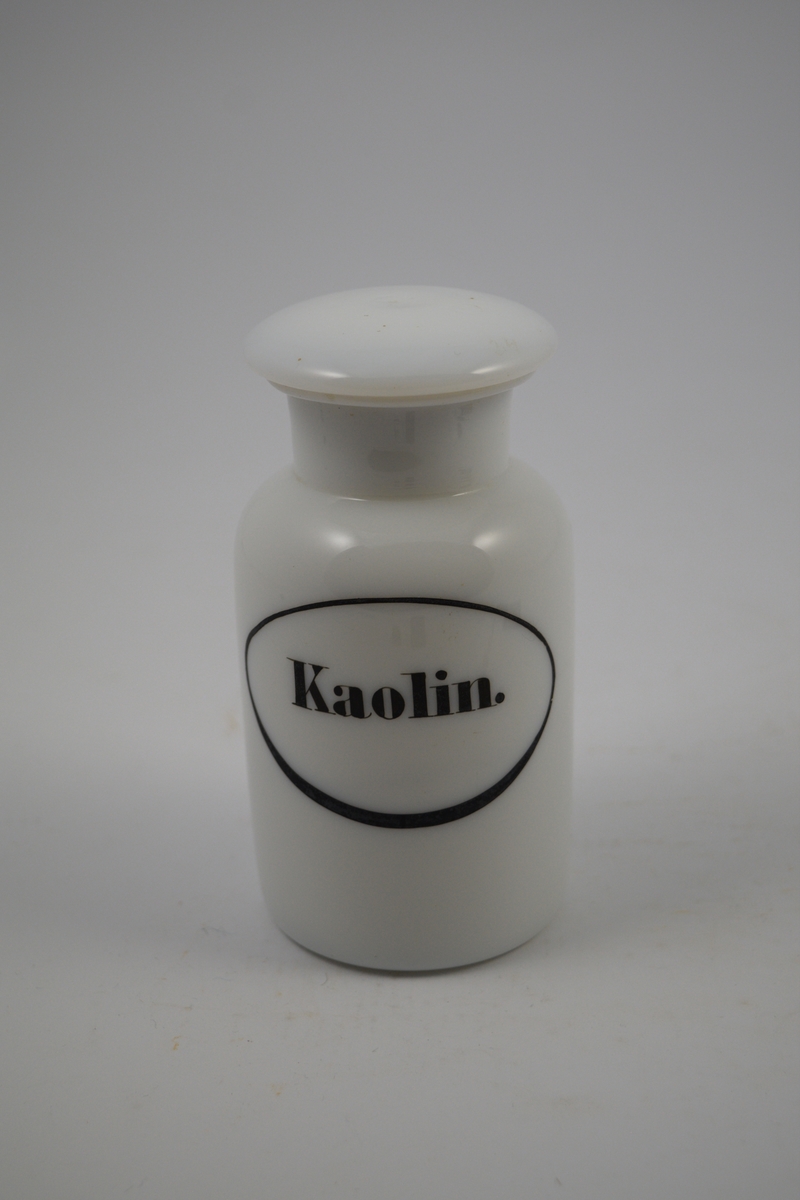 Hvit porselenskrukke med hvit propp. Påsatt etikett med sort oval ramme og sort skrift. Kaolin er syrenøytraliserende og ble brukt mot mageproblemer.