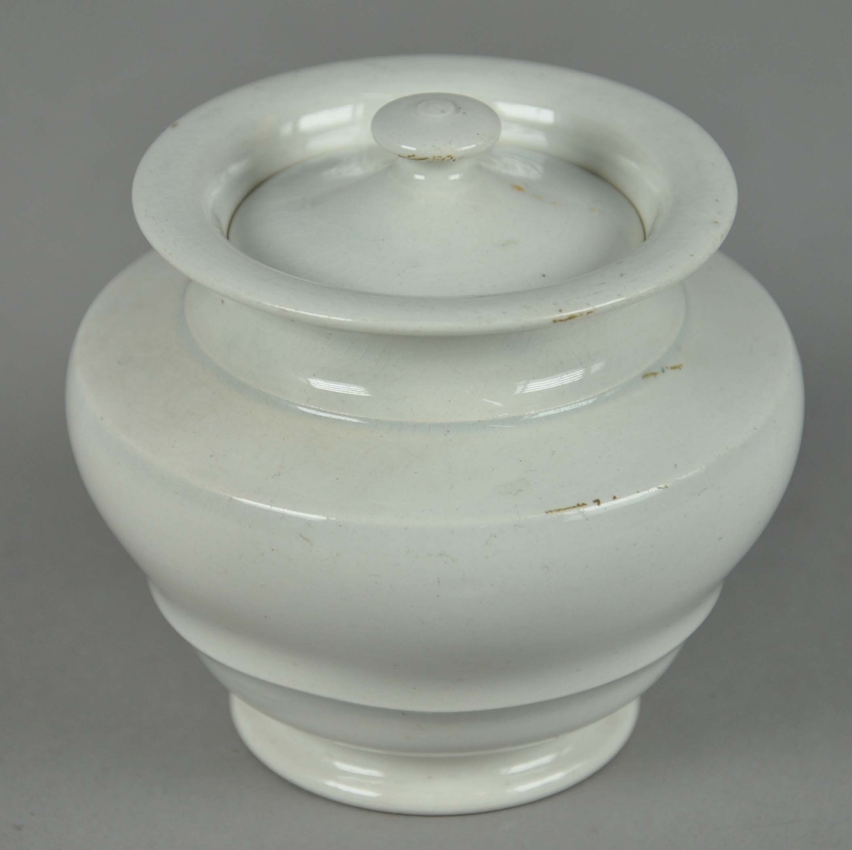 Sukkerskål av glassert keramikk, med lokk. Skålen har en rund form med utsående korpus, med profilert snitt.