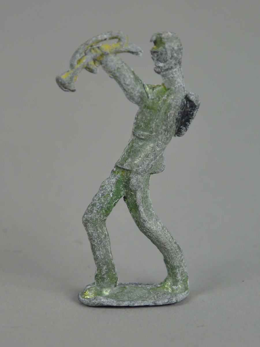 Liten figur av tinn. Figuren er i marsjposisjon på en oval støtte og blåser i trompet.
