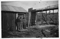 Tre arbeidere står foran ei brakke. Kalkovn i bakgrunnen. Lu