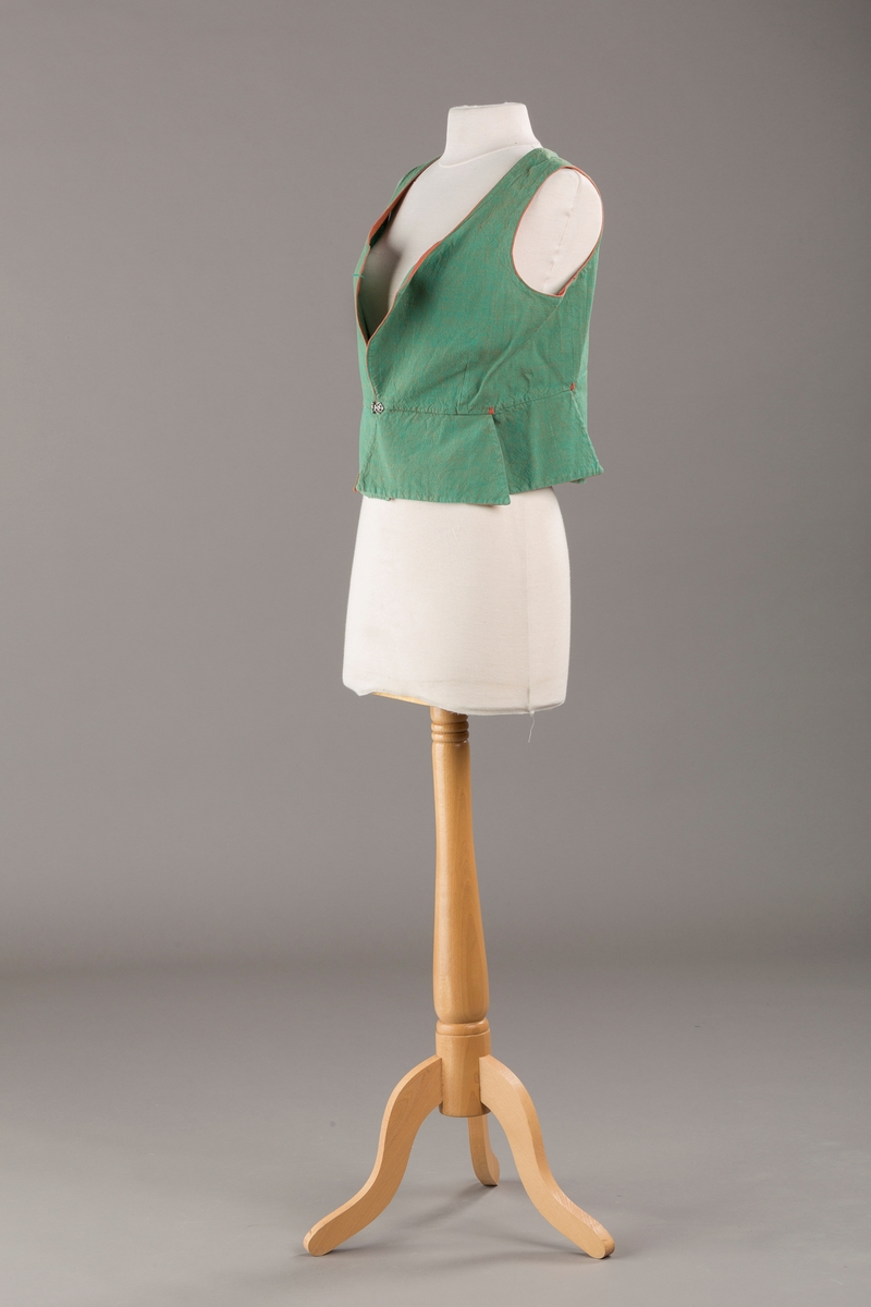 Grønt liv med rustrød kant og fór, seks skjøter. Håndsydd forsterkning i kontrastfarge øverst i skjøtesplittene. Lukkes med tinnhekte.