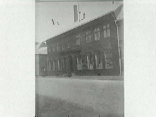 Tegelhus med skylt för "Martin Svensson, ... Falck & Co", i stadsmiljö. I bakgrunden syns en skorsten och till vänster i bild promenerar två kvinnor.