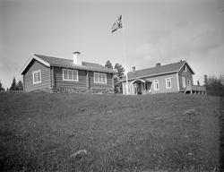 To hus, det ene i tømmer,  med  flaggstang i mellom.