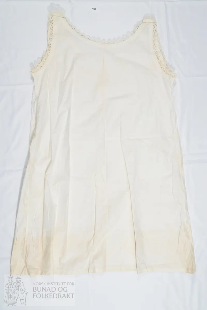 Nattkjole i bomullslerret med smal heklet kant rundt hals og ermringing. Sydd av et for- og bakstykke, med knepping med knapphull og knapp på skuldrene. Sydd sammen med indresøm på maskin. Svært små sting. Den påsydde blonden er heklet av mercerisert bomullsgarn. Dobbel oppbrett nederst på skjørtekanten.