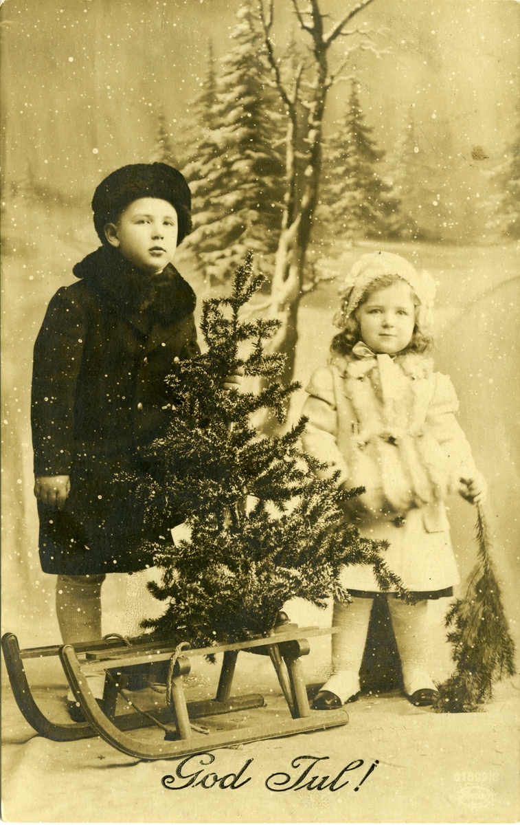 Julekort. Jule- og nyttårshilsen. Fotografisk motiv. Svart/hvitt. Vinterlandskap. En gutt og en jente står ved siden av en kjelke. På kjelken står et juletre.