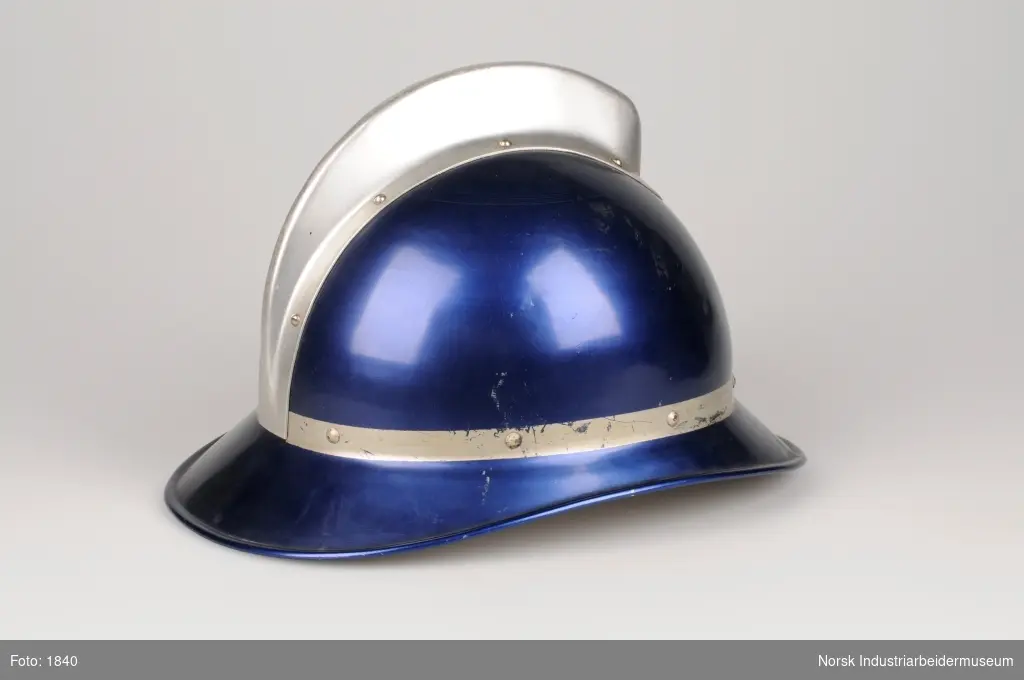 Blå brannhjelm i metall. Sølvfarget rim rundt hjelm og sølvfarget topp. Indrehette av skinn og festereim i skinn med metallspenne.