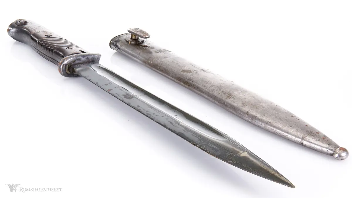 Metallhåndtak med lang pommel og geværfeste med fjæret knapp i bakkant. Serratert tregrep som er festet med 2 skruer. Langt spisst stålblad med egg og blodrand. Metallslire med kule enden og krok for festing til grunnutrustning/belte.