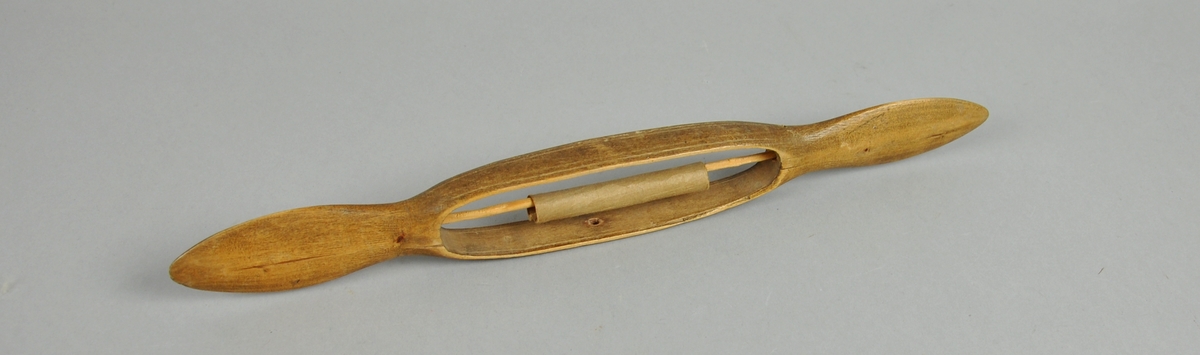 Avlang vevskyttel med flate håndtak i hver ende. Det er skåret ut et hull på midten med spile som det er surret et papir rundt.