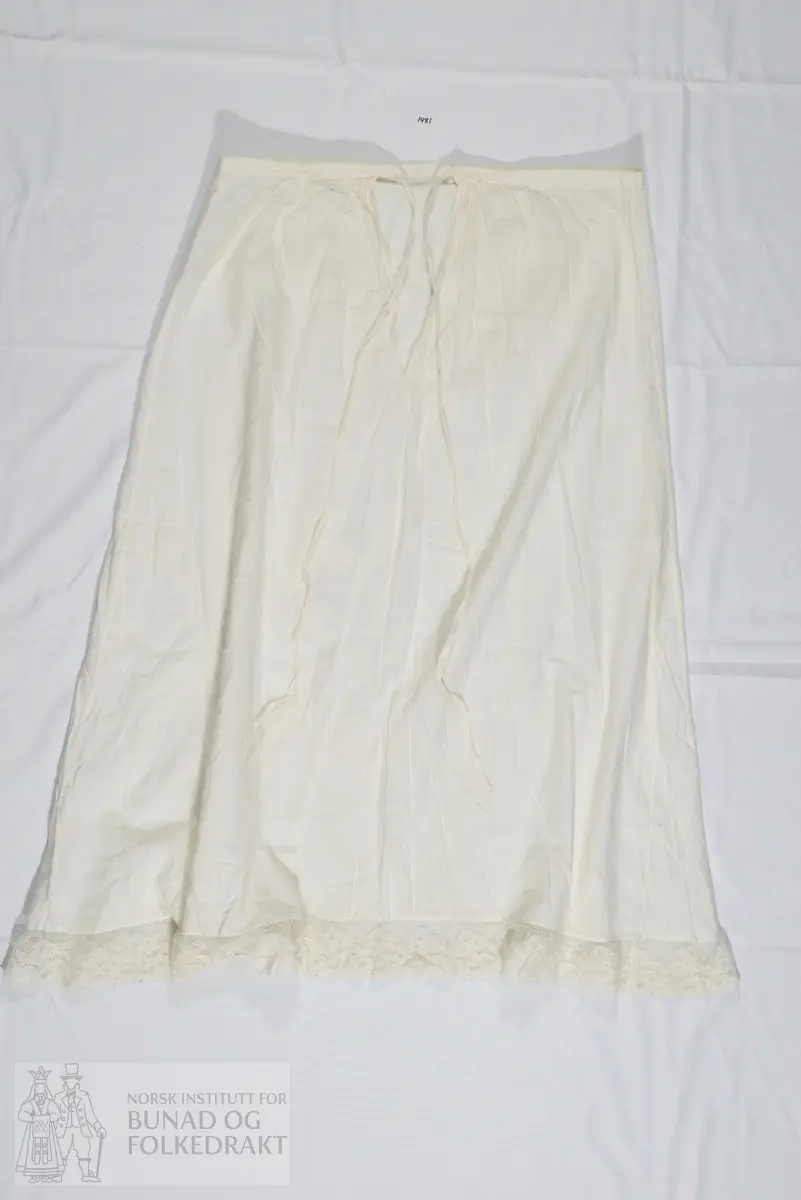 Understakk i hvit bomull. Kantet nederst med kjøpeblonde som trolig også er av bomull. Understakken har en smal linning øverst og lukkes med knytting. Sydd sammen av to høyder stoff som er lagt i små rynker til linningen. Ved linningen finner vi også to små innsnitt.  Understakken har i tillegg 2 isydde kiler fra omtrent knehøyde, disse er noe ujevne og har ikke helt samme høyde. Linningen har en skjøt midt bak. Maskinsøm