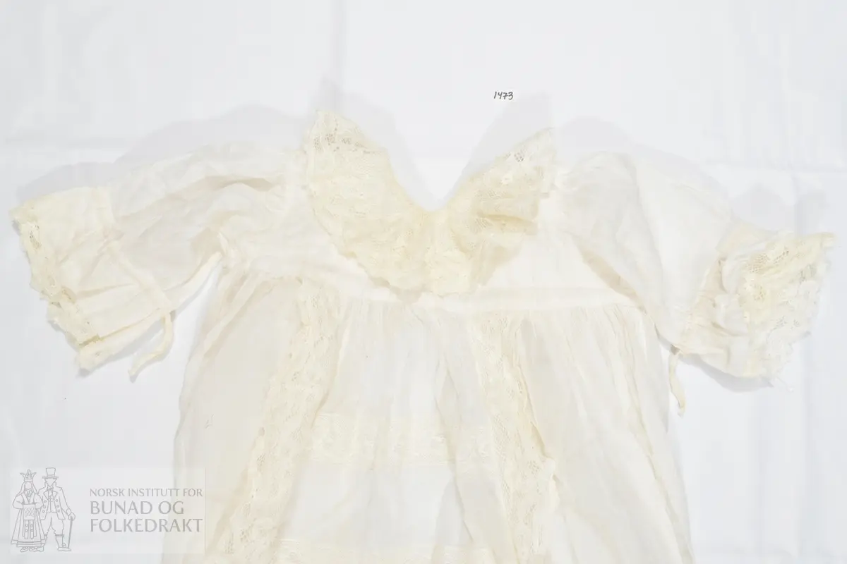 Lang dåpskjole i hvit bomullslerret med påsydde kjøpeblonder. Livstykket er sydd av to lag med tynn bomullslerret. Sydd til skjørtet med en bred linning. Ermer sydd rett i, med rynker langs hele ermeåpningen. 

Åpning midt bak med splitt et stykke ned på skjørtet, knyting øverst med bånd. Forstykke og de to bakstykkene er sydd sammen på skuldrene. Halslinning er brettet ned dobbelt på baksiden, og er sydd ned på maskin. Bred blonde i halsringingen som er sydd på med maskin. Blonden er maskinlaget, og er av bomull. Den er noe rynket til halslinningen. 

Ermet er sydd av et enkelt lag med tynn bomullslerret, og har påsydd en løpegang for knytesnorer på vrangen. På vrangen er også festet en bred blonde som stikker ut av ermeåpningen.

Skjørtet er tett rynket til linningen i livet og sydd ned med maskin. Skjørtet er sydd av fem høyder, sydd sammen med maskin. Et felt midt foran er dekorert med blonder langs skjørtehøydene på hver side, og som går hele veien ned til skjørtekanten. I tillegg er horisontale blondebånd sydd inn mellom felt av bomullstoffet i skjørtehøyden midt foran. Alt er sydd på maskin. Nederst på skjørtet er det påsydd en bred kappe, som er lett rynket til skjørtet. Kappen har mellomverk av maskinblonde.