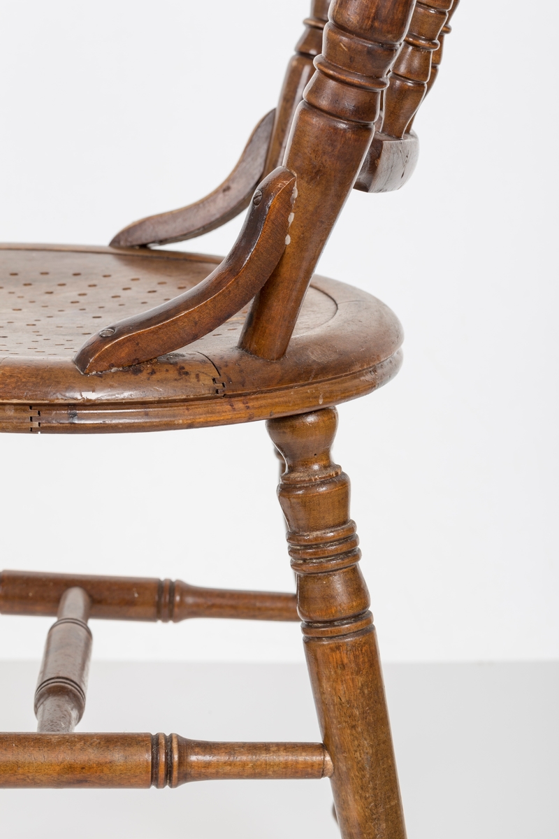 Stol i lakket treverk. Rundt stolsete med hullemønster. Rygg som er svakt buet med spiler og mønster. Dreide ben om spiler i rygg.