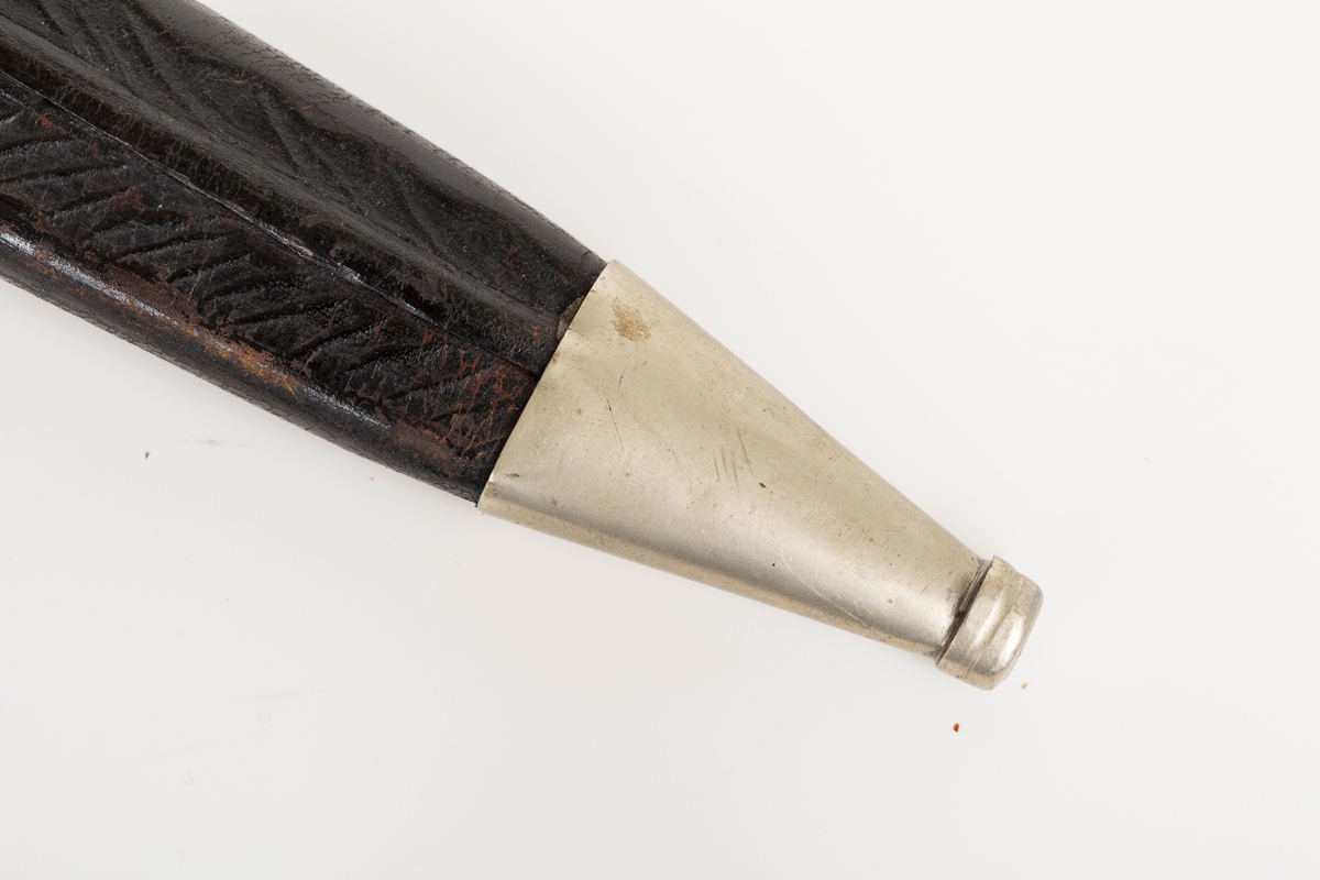 Håndsydd lærslire med holker oppe og nede. Slire og stropp av separate lærstykker. Sliren har enkel  pautet dekor. Holkene er håndloddet nysølv (sansynligvis) med enkel trambulert dekor. Toppholken har påloddet hengsle og D-ring til feste for slirestropp.