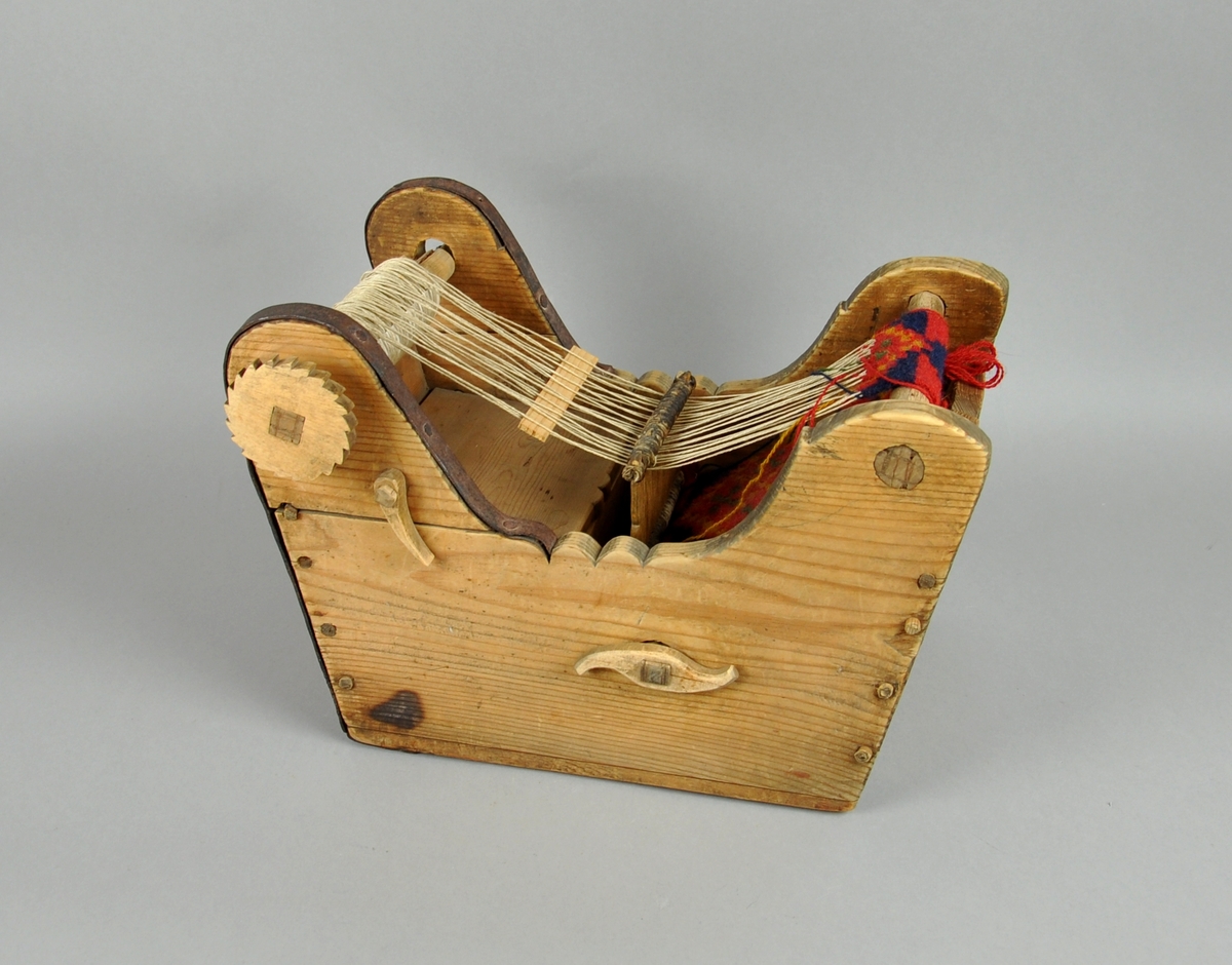 Trekasse med brystbom, garnbom og slagbom. Veven har renning og det er vevet et lite stykke bånd med mønster i rødt, blått og grønt.