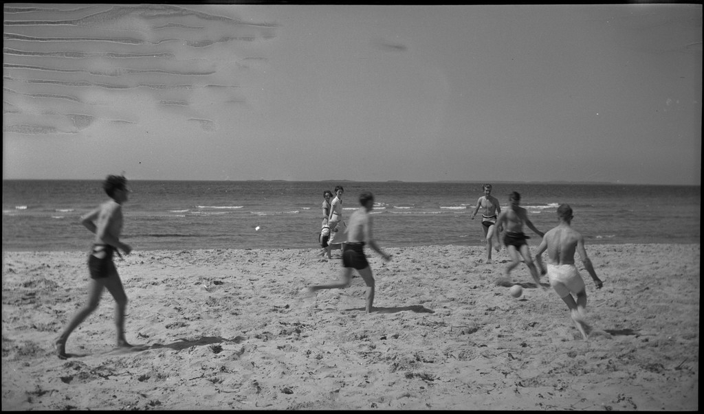 En stor vennegjeng i lystig lag på stranda på Sola. De har trommer og trekkspill og leker i vannet og på land med og uten ball. Sola strandhotell ligger i bakgrunnen. Tor Johannessen og Harald Bergsaker er på bilde nr. 14 og 18.