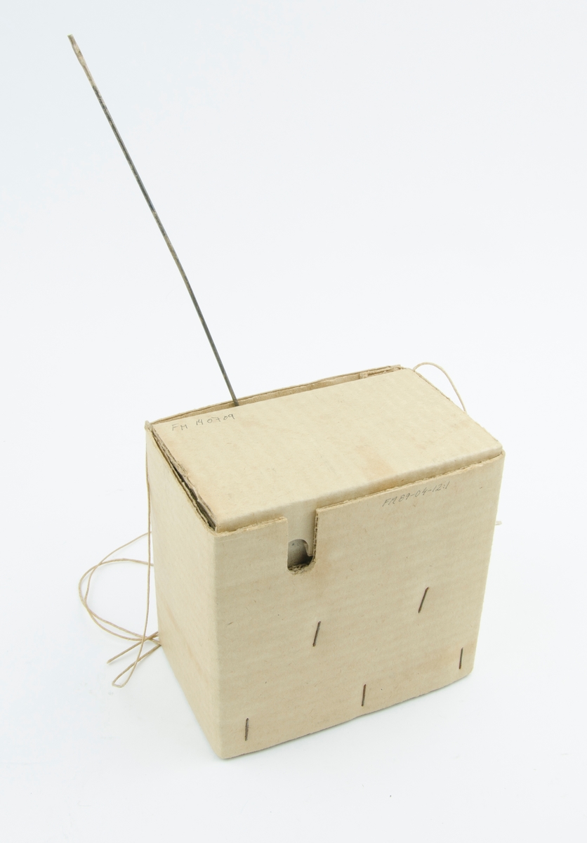 Papplåda med fyra snören och en antenn. I lådan finns utrymme för sändaren. Pejlsändaren har troligen en funktion inom höjdmätning.
