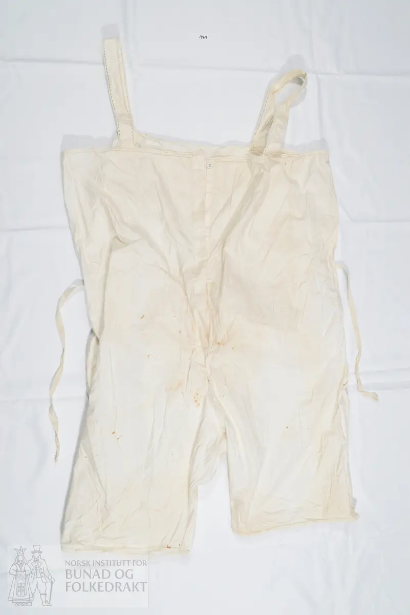 Chemilette, underbukse med overdel/ linnet, i hvit bomull. Knelang bukse, overdel med seler og åpning midt framme med knepping. Splitt i sidene og horisontal åpning bak. Overdelens ryggstykke stanser ved livlinjen. Lokk bak på buksen, med knytesnor øverst på lokket i hvert hjørne. Knytebånd til å knyte rundt livet. Pen avslutning på benene, med sydd hullfald og leggsøm, samt en smal heklekant helt ytterst.