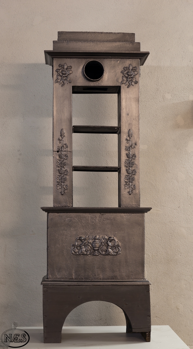 Ovnen består av to avdelinger. Underdelen har et frembuet midtparti med kamskjell flankert av sjødyr på hver side. På kortsidene skjold, løvehode og symmetriske ørner. På bakplate en krukke med symmetriske engler.
Overdelen er høy, firkantet med tredelt åpning. Empire ornamentikk og avtrappet topp. Øverst på frontplaten symetriske fabeldyr (pegasus) vendt mot hverandre.