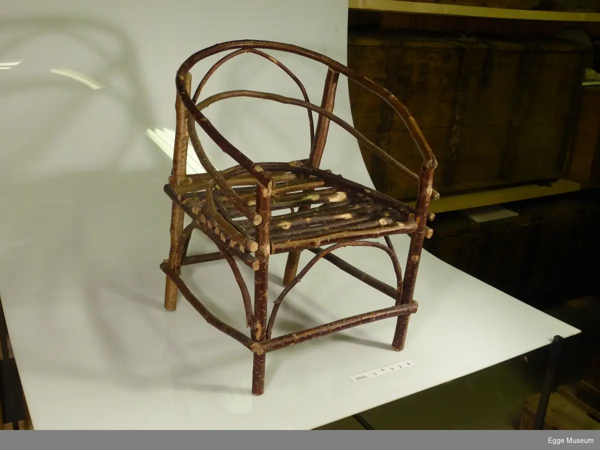 Barnestol konstruert av trekvister av ulike løvtresorter som er spikret sammen. Form: firkantet sete med to buede støttekvister. Fire bein. Buet rygg som går over i armlener. Stolen ble laget under krigen.