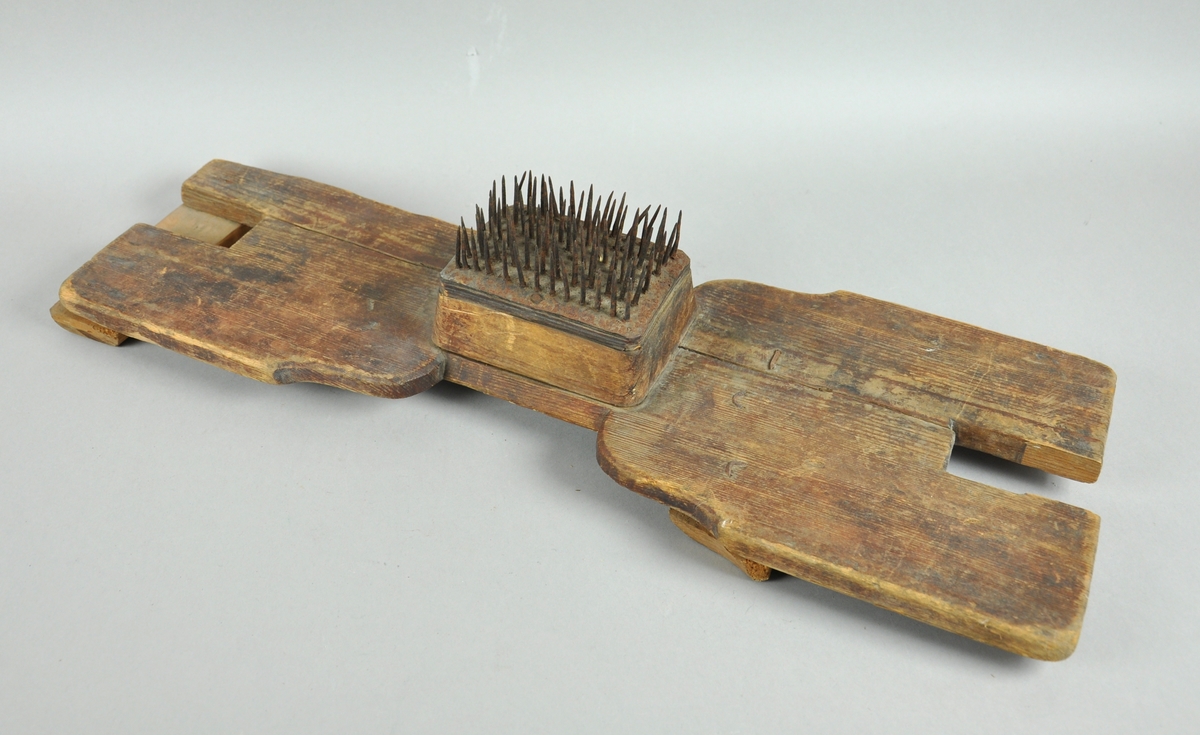 Linhekle av tre som er smalere på midten, der det er festet en trekloss med stålspiker. Trebrettet er skadet og reparert ved å spikre på to trestykker på tvers på undersiden.