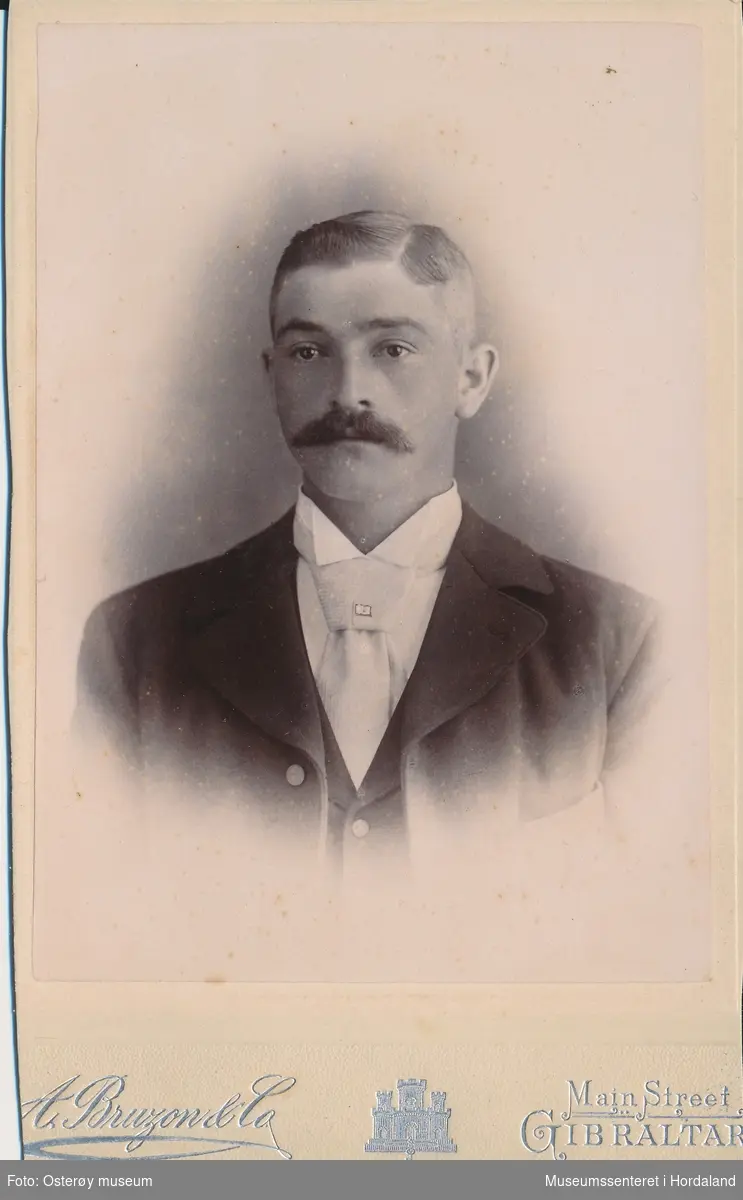 atelierfotografi i halvfigur av ung mann med bart, mørk jakke, vest, kvit skjorte og ljost slips