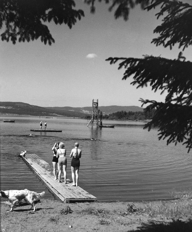 Svart-hvitt foto av badebrygge med tre barn som står klare til å hoppe i vannet i Mjøsa, og et stupetårn av tre lenger ut i vannet.