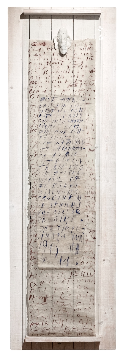 Objekt, "Bikt" av Kerstin Suni. På platta av vitmålade bräder är hängt en smal och en bred rispappersremsa med text i blått resp. brunt. Överst ett stiliserat ansikte i gips. Allt täckt med en glasskiva.