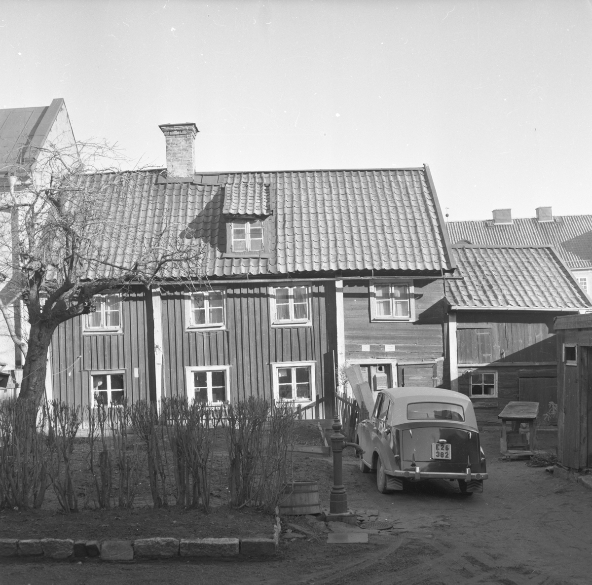 Motiv från Hunnebergsgatan 25 i Linköping 1961. Boningshuset med bod och uthus sedda från gården. Bilen är en Triumf Mayflower från 1951.