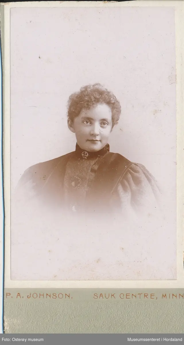 portrettfotografi av ung kvinne med krølla hår satt opp, vid mørk jakke med store, rynka ermer, svart band om halsen med nål
