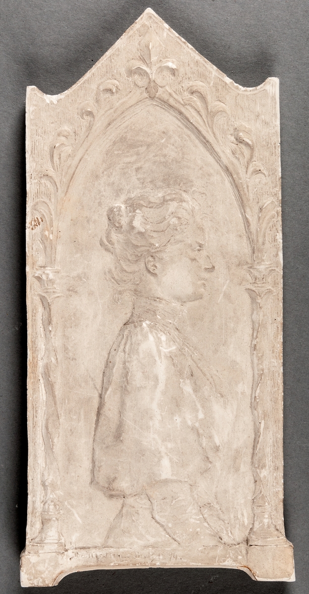 Gipsrelief föreställande kvinna i profil. Omgivande dekor i nyrennässansstil.
Signerad Ida Matton Paris 1894.
