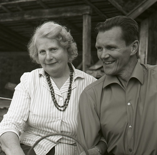 Leende Lisa Viksten och Hilding sitter armkrok vid ett bord utomhus framför en träbyggnad, 19 augusti 1962.