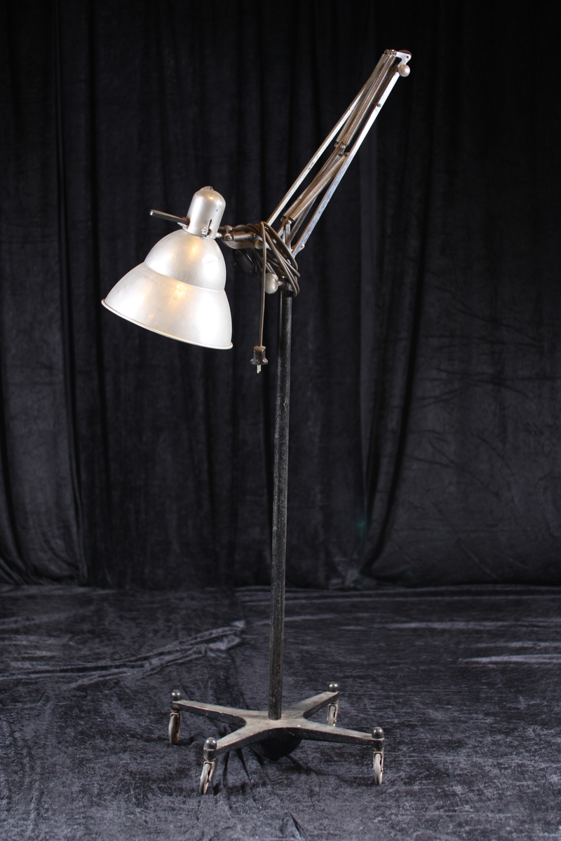 Regulerbar lampe nytta i fotoatelier til belysning av motiv. Lampen står på ein tung støypejernfot med fire armar og hjul.