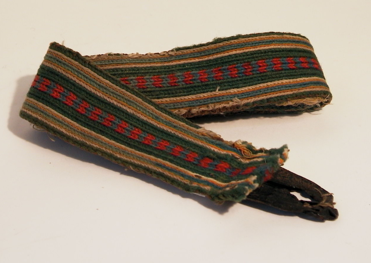 Hängselband, brickat eller slånnat med ullgarn i rött och grönt, brunt och blått samt oblekt lingarn. Knapphål av läder.