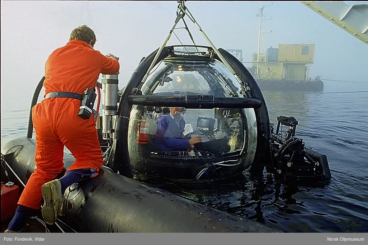 En mann står i en gummibåt og jobber med noe på miniubåten "Check Mate" som henger i kranen på moderfartøyet "Strilborg".  To menn sitter i ubåten. I bakgrunnen ligger dykkerlekteren "Buldra".