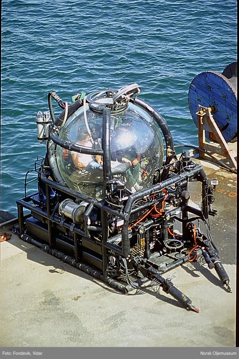 To personer står ved siden av miniubåten "Check Mate", mens to andre personer sitter inne i ubåten.