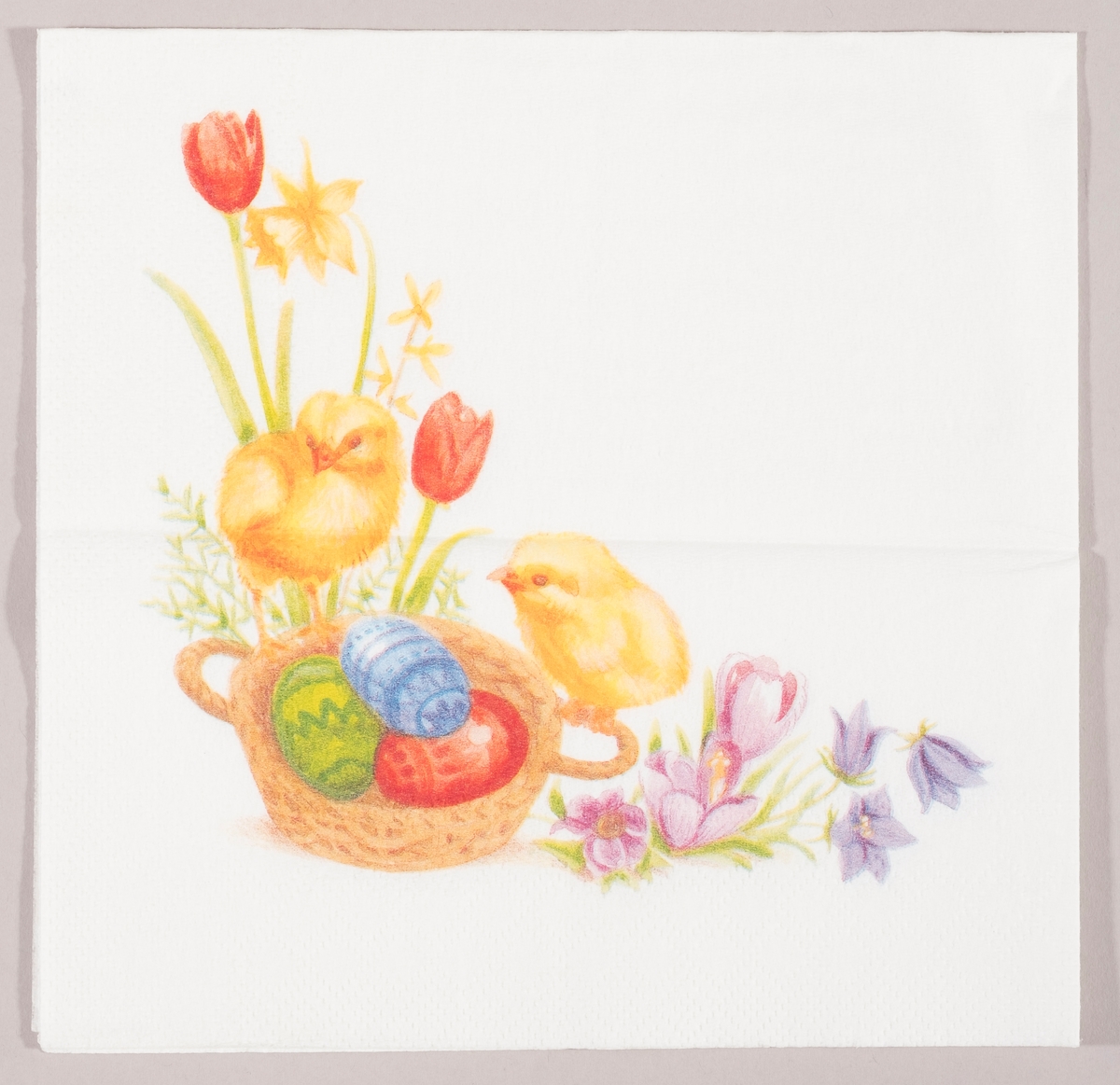 To kyllinger sitter på en flettet kurv med dekorerte påskeegg. Rundt om kurven er det tulipaner, påskeliljer, krokus og blåklokker.