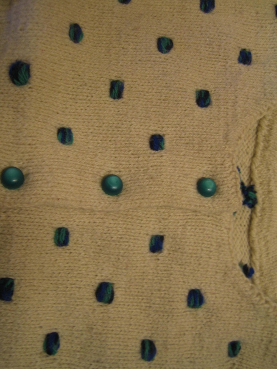Kofta i naturvit ull slätstickad med invikta kanter i märmslut, halskant och nederkant. Koftan har sex knapphål och knappar i turkos. Koftan är dekorerad med blågröna prickar av ullgarn som är knutna på avigsidan med ett flätat avslut.