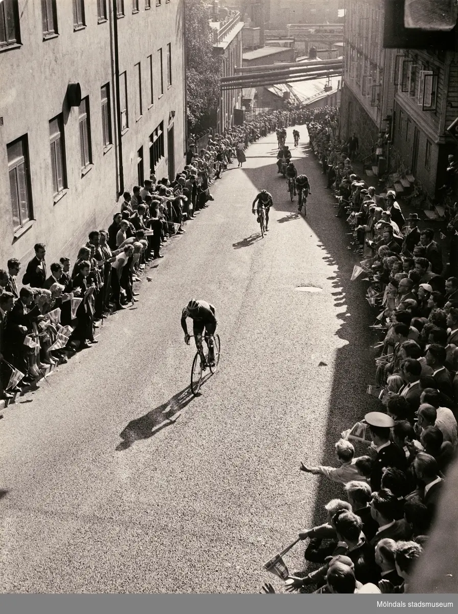 Cykel-SM söndagen den 5 augusti 1956 i Mölndal. Tävlingscyklister tar sig uppför "Kråkan" (Kvarnbygatan) under seniorernas 18-milslopp. Backen kantas av stor publik på båda sidorna. Starten gick klockan 12 och inleddes med en varvbana inom Mölndal på följande gator: Järnvägsgatan-Kvarnbygatan-Stensjögatan-Muraregatan-Gunnebogatan-Järnvägsgatan. Varvbanan kördes tre gånger varefter cyklisterna åkte landsvägsslingan på cirka 13 mil över Landvetter-Härryda-Hällingsjö-Älvsåker-Ubbhult-Hällingsjö-Inseros-Eskilsby-Härryda-Mölnlycke och när de åter kom till Mölndal åktes ovan nämnda varvbana åtta gånger till. Segrade gjorde Roland Ströhm före tvåan Karl-Ivar Andersson och Lars Nordwall på bronsplats.
Relaterat motiv: 2023_0153
