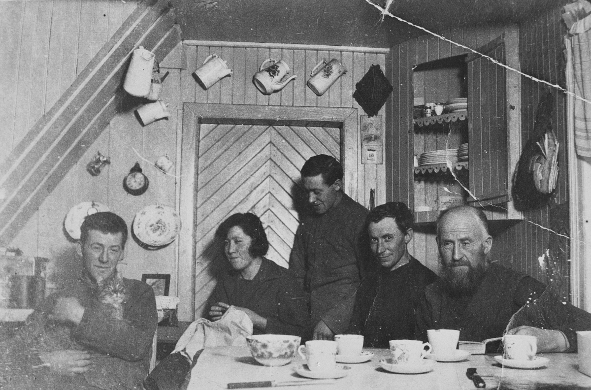 Gruppebilde innendørs, 4 menn, 1 kvinne og 1 katt samlet rundt et dekket bord, ca. 1920.