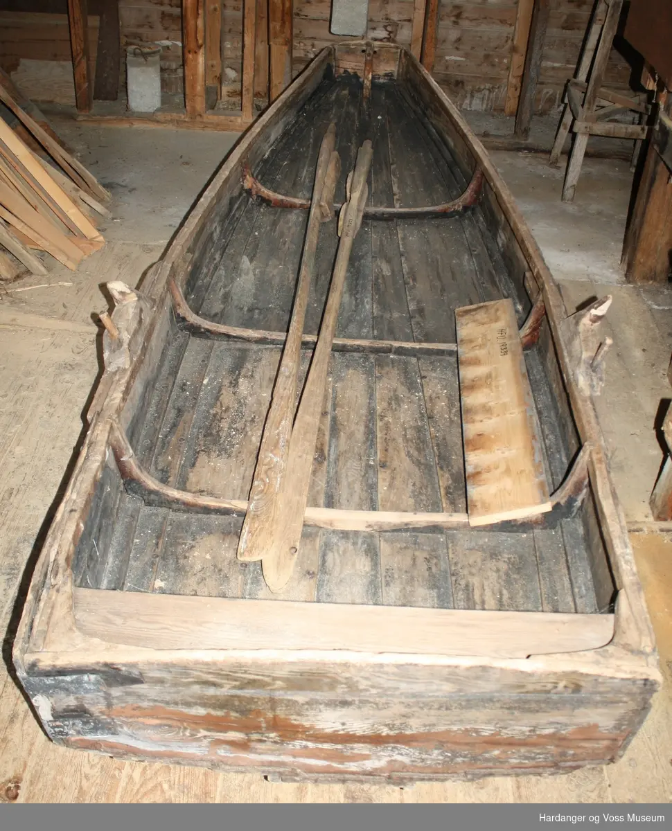 Klinkbygget pram av furu. Båten har en løs tofte og et par keiper med tollepinner. Tilhørende båten er et par årer, også av furu.